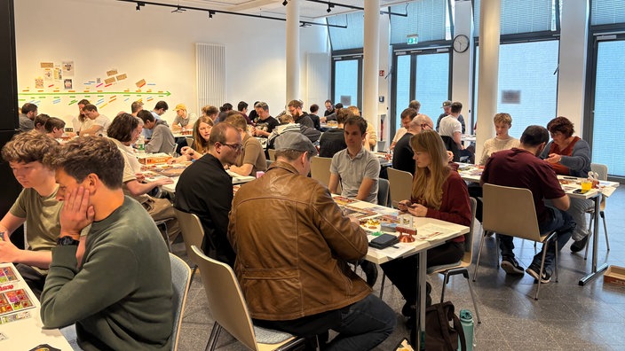 An zahlreichen Tischen sitzen Menschen und spielen ein Brettspiel