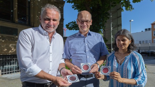Drei Personen, die Aufkleber präsentieren. Links Thomas Köhler. Daneben Prof. Dr. Rainer Stamm und  Dr. Anjali Scholten.