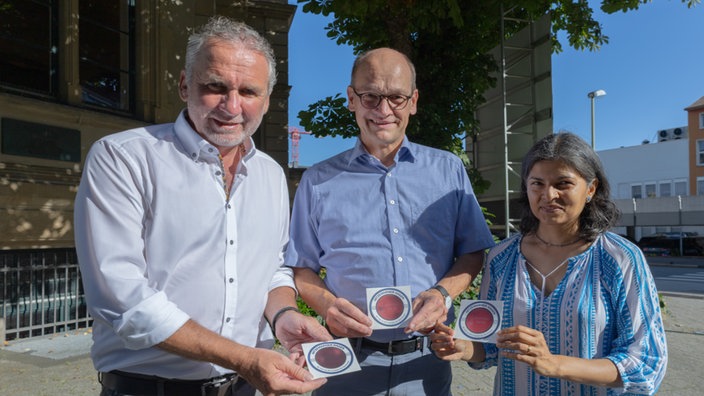 Drei Personen, die Aufkleber präsentieren. Links Thomas Köhler. Daneben Prof. Dr. Rainer Stamm und  Dr. Anjali Scholten.