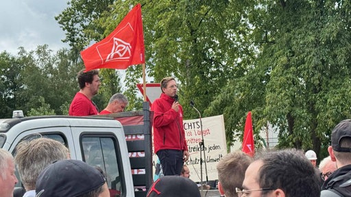 Drei Personen in roter Kleidung stehen auf einem Fahrzeug und sprechen zu einer Menschenmenge
