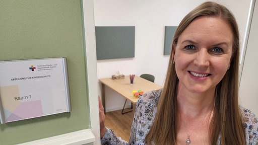 Frau steht auf der rechten Seite in einem Türrahmen und links ist ein Schild für die Raumbeschreibung 
