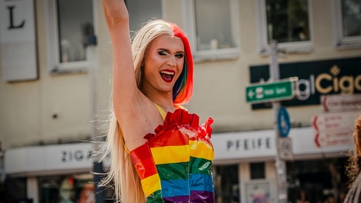 Eine Person in einem Regenbogenkleid aus Kunststoff reckt einen Arm in die Luft und zwinkert in die Kamera. Sie ist stark geschminkt und hat lange Haare. Eine Hälfte blond, die andere rot und grün. Es ist optisch eine Frau.