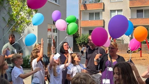 Mehrere Schüler und Erwachsene stehen mit Helium-Ballons mit Karten daran auf einem Hof.
