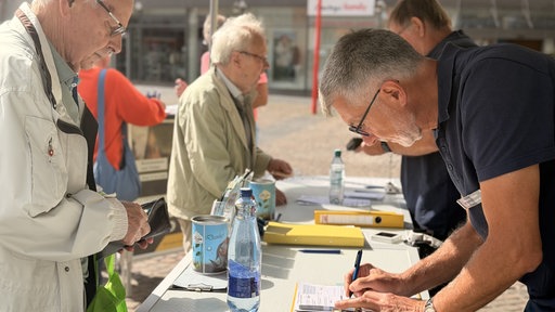 Mehrere Männer stehen an einem Tisch auf einem Marktplatz, um Wasserproben aus ihren Brunnen abzugeben.