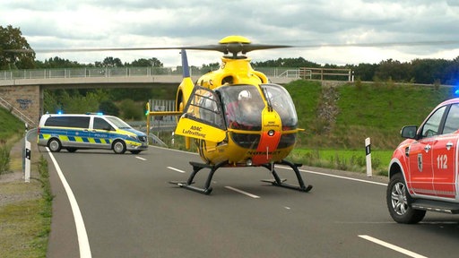 Ein gelber ADAC-Hubschrauber, der auf einer Straße gelandet ist, daneben Feuerwehr und Polizei.