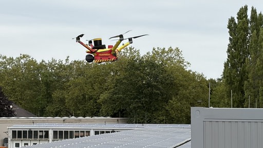 Eine Drohne in Feuerwehrfarben schwebt vor Bäumen.