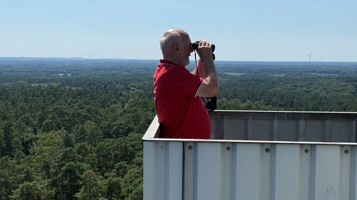 Ein Mann steht mit einem Ferngals auf einem Turm im Wald 