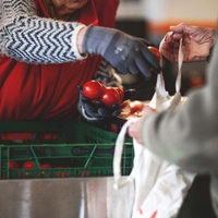 Symbolbild: eine alte Damen reicht einem Mann Tomaten über grüne Kisten hinweg.