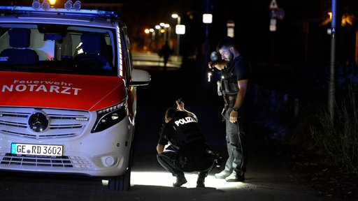 Zwei Polizeibeamte suchen mit der Taschenlampe auf dem Boden nach Spuren, daneben steht ein Rettungswagen. Es ist Nacht. 