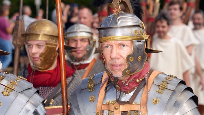 Ein Mann mit Helm und Rüstung steht inmitten anderer Männer. 