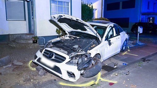 Eine stark beschädigtes Auto mit offener Motorhaube, ausgelösten Airbags steht zur Hälfte in einem Vorgarten.