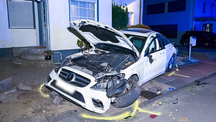 Eine stark beschädigtes Auto mit offener Motorhaube, ausgelösten Airbags steht zur Hälfte in einem Vorgarten.