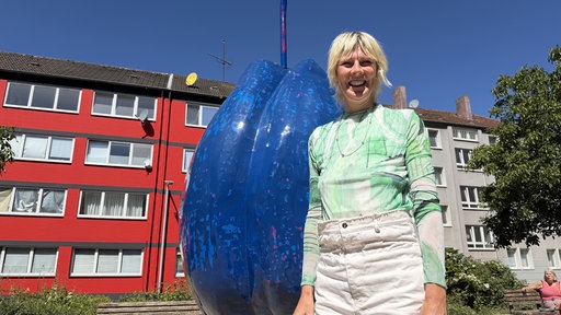 Eine pflaumenförmie Skulptur auf einem Platz, neben ihr steht die Künstlerin - lachend, mit blonden Haaren.