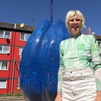 Eine pflaumenförmie Skulptur auf einem Platz, neben ihr steht die Künstlerin - lachend, mit blonden Haaren.