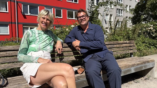 Eine blonde Ferau und ein dunkelhaariger Mann mit Brille sitzen auf einer Holzbank.