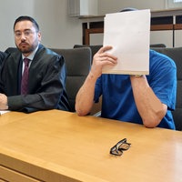 Ein Mann mit blauem Shirt sitzt in einem Gerichtssal und hält sich Papier vor das Gesicht. Daneben sitzen ein Mann und eine Frau in Robe.
