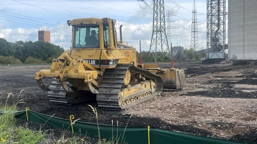 Ein gelber Bagger steht vor einer Baustelle, im Hintergrund stehen viele Hochspannungsmasten. 