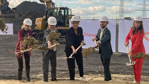 Fünf Menschen stehen mit weißen Helmen auf einer Baustelle und schaufeln Erde, ein feierlicher Festakt. 