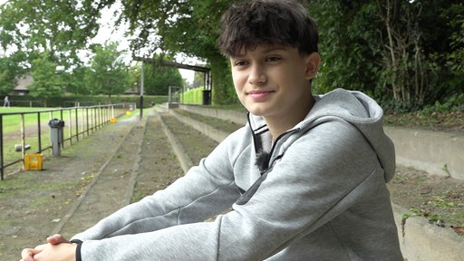 Ein Teenager mit dunklen Haaren, gekleidet mit einem grauen Kapuzenpullover, sitzt auf einer Tribüne und lächelt in die Kamera