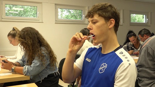 Im Vordergrund ist ein junger Mann in einem blau-weißen Sporttrikot zu sehen. Er hält ein Wattestäbchen in der Hand und führt es in den Mund ein, um einen Abstrich aus der Mundschleimhaut zu nehmen.