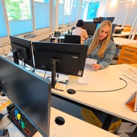 Eine blonde junge Frau sitzt in einem Büro am Schreibtisch.
