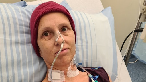 Maria Fischer liegtmit einer Mütze auf dem Kopf und einem Schlauch in der Nase in einem Krankenhausbett.