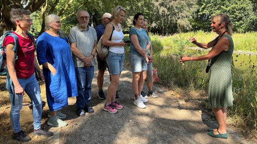 Ursula Stratmann in grünem Kleid steht im Wald und erklärt etwas einer Gruppe Menschen 