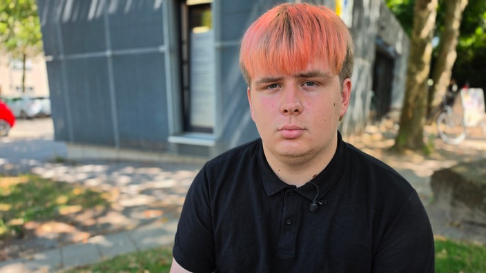 Ein junger Mann guckt verärgert in die Kamera. Er hat ein rundes Gesicht und pink gefärbte Haare.