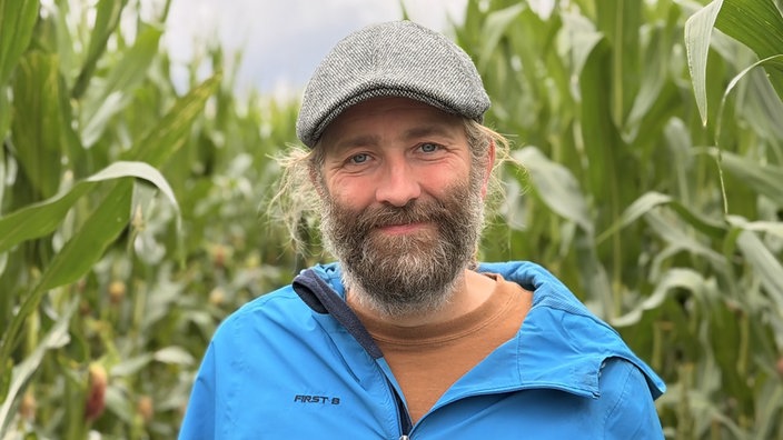 Ein Mann mit Vollbart und grauer Mütze steht vor Maispflanzen.