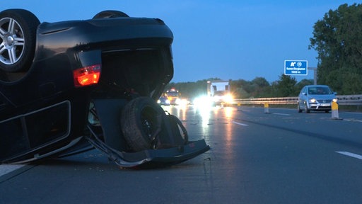 Ein PKW liegt auf einer Autobahn auf dem Dach, die Kofferraumklappe ist offen. Im Hintergrund sind andere Autos auf der Fahrbahn zu sehen. 