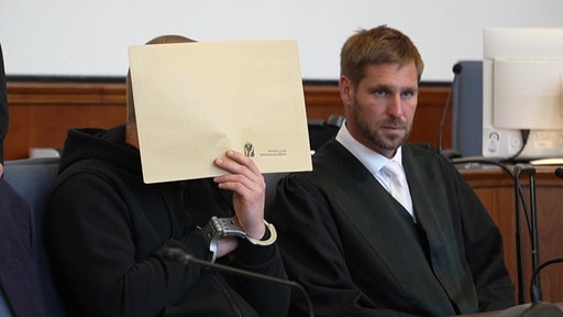 Der Angeklagte hält sich im Gerichtssaal eine Aktenmappe vors Gesicht
