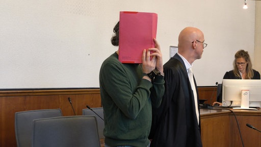 Der Beschuldigte verdeckt sein Gesicht mit einer Mappe. 
