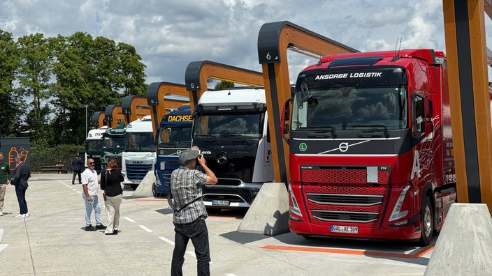 Sechs LKW stehen an den jeweiligen Ladesäulen im neuen Ladepark, davor stehen Personen.