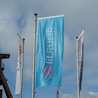Zwei wehende blaue Fahnen mit der Aufschrift "Lit.Ruhr" stehen neben dem Förderturm der Zeche Zollverein in Essen