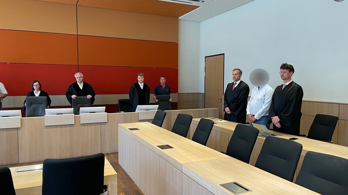 Der Angeklagte, seine Verteidiger und weitere Mitarbeitende in dem Gerichtssaal.