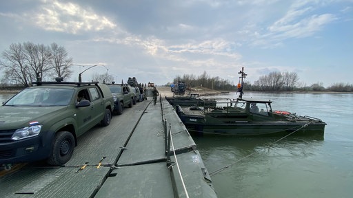 Mehrere Militärfahrzeuge parken an einem Steg, an dem ein Militärboot liegt.