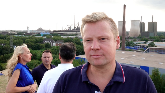 Sebastian Paradis spricht während eines Interviews. Im Hintergrund stehen Jochen Taczkowski, Diana Leenen und Hrvoje Vlaovic sowie die Industriekulisse im Duisburger Norden.
