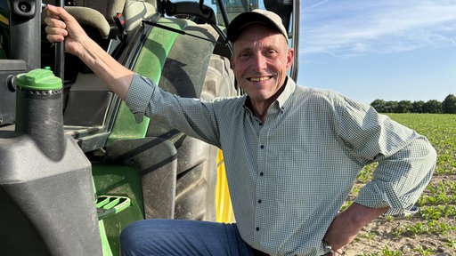 Ein Mann hält sich an seinem Traktor fest und lacht gut gelaunt in die Kamera. Der Traktor steht auf einem Feld