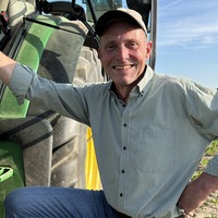 Ein Mann hält sich an seinem Traktor fest und lacht gut gelaunt in die Kamera. Der Traktor steht auf einem Feld