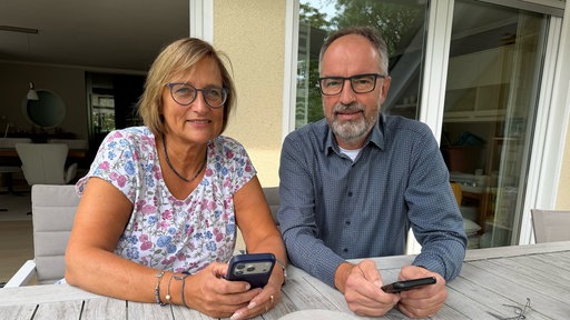Claudia und Uwe Brock sitzen am Gartentisch mit ihren Smartphones in der Hand.