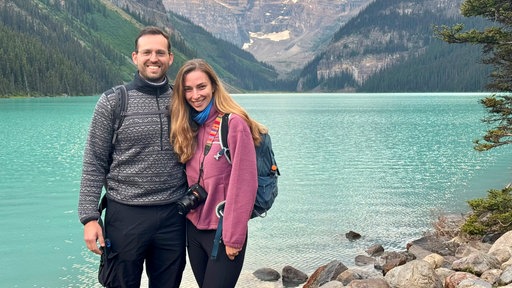 Alexandra und Lukas stehen vor einem großen See mit klarem, blauem Wasser.