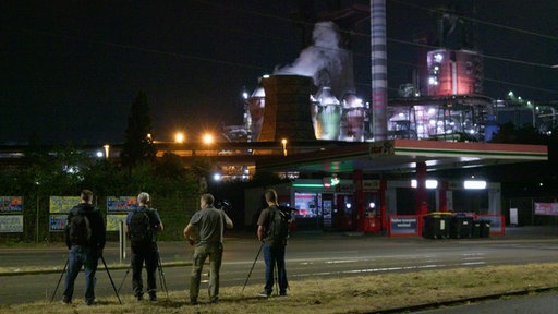 Teilnehmende stehen mit ihren Kameras vor einer Tankstelle, im Hintergrund Industrieanlagen 