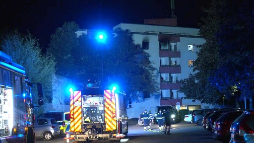Eine Wohnanlage mit verrußtem Fenster und Balkon bei Nacht, davor ein Feuerwehrauto mit Blaulicht.