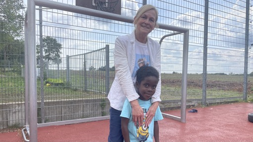 Claudia Peppmüller vom Friedensdorf International und Amari stehen zusammen auf dem Fußballfeld vor einem Tor.
