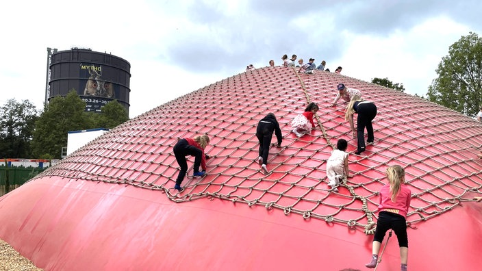 Kinder klettern auf einem großen roten Kissen, im Hintergrund der Gasometer