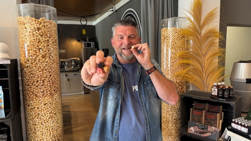 Michael Althoff hält mehrere Popcornstücke in die Kamera und isst eines, im Hintergrund große Behälter mit Popcorn.
