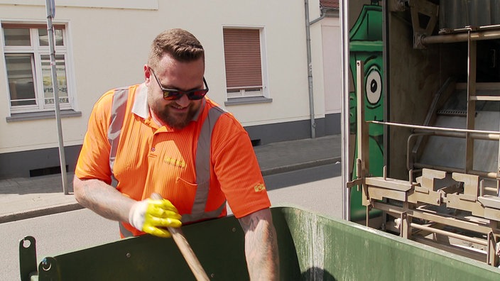 Das Bild zeigt Max Müller dabei, wie er einen Müllcontainer mit einer Schaufel reinigt.