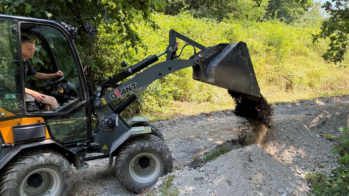 Ein gelber Bagger mitten auf einem schmalen Waldweg schafft eine Schaufel mit Erde an die Seite.