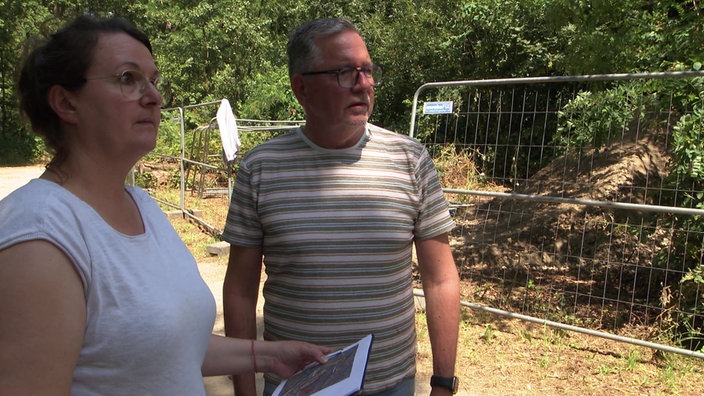 Eine Frau und ein Mann stehen vor der Baustelle im Wald und überwachen die Arbeiten.