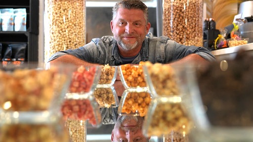 Michael Althoff sitzt hinter verschiedenen Popcornsorten in kleinen Schalen und blickt in die Kamera, während im Hintergrund große Popcornbehälter stehen.
